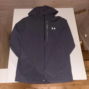 Mörkgrå vindjacka från Under Armour - Snygg svart vindjacka från Under Armour med huva och hel dragkedja framtill. Jackan har logga och text på bröstet, samt diskreta fickor i sidorna. Perfekt för dig som gillar sportig stil och vill ha något lätt och funktionellt. (OBS dragkedjan är trasig men det går forfarande att knäppa upp och ner jackan felfritt, går förmodligen och fixa dragkedjan hos någon kunnig‼️‼️)