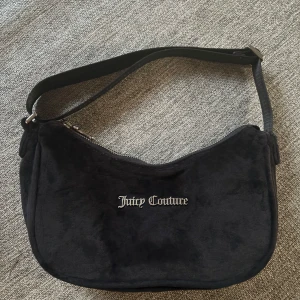 Svart axelväska från Juicy Couture - Snygg svart axelväska från Juicy Couture i mjuk sammet med silverfärgad logga framtill. Väskan har en enkel, stilren form och stängs med dragkedja upptill. Perfekt för dig som gillar klassisk Y2K-stil och vill ha en ikonisk accessoar.