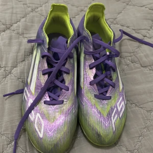 Adidas F50 lila fotbollsskor elite - Säljer ett par Adidas F50 fotbollsskor i lila och neongult med vita detaljer. Skorna har snörning, låg profil och mönstrad ovandel i syntetmaterial. Yttersulan är delvis transparent och neongul med fasta dobbar för bra grepp på planen. Perfekta för dig som vill sticka ut på matchen!