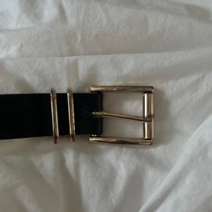 Svart skärp med guldspänne - Stilrent svart bälte med rektangulärt guldspänne och två matchande guldfärgade hällor. Perfekt accessoar för att lyfta din outfit med en klassisk touch. Bältet är i slätt material och har en enkel, tidlös design.