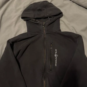 Svart hoodiejacka från Sail Racing - Svart hoodiejacka från Sail Racing med dragkedja framtill och bröstficka med logga. Jackan har huva med snörning och en sportig look, perfekt för dig som gillar stilrena och funktionella plagg.