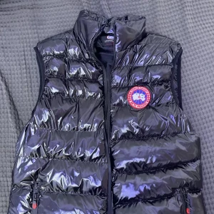 Svart dunväst från Canada Goose - Säljer en glansig svart dunväst från Canada Goose med klassisk logga på bröstet. Västen har hög krage, två dragkedjefickor och quiltad design. Perfekt för lager-på-lager och riktigt snygg till streetwear eller chill outfits. Köptes för 3 månader sen men har använt den kanske 5-6 gånger för storleken passar inte mig längre. Köptes 2000kr. Pris kan diskuteras!