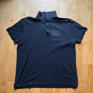 Svart pikétröja Peak Performance XXL - Svart pikétröja från Peak Performance i storlek XXL. Tröjan har korta ärmar, ribbad krage med tre knappar och tryckt logga i flera rader på bröstet. Materialet är mjuk bomull som känns skönt mot huden. Perfekt för dig som gillar stilren och sportig look.