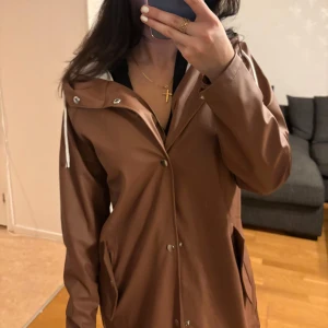 Regnjacka med huva och knappar - Snygg regnjacka med unik färg och med huva och vita dragsnören. Jackan har tryckknappar framtill, två fickor med lock och en längre, rak passform. Perfekt för regniga dagar och enkel att matcha med olika outfits.🤍