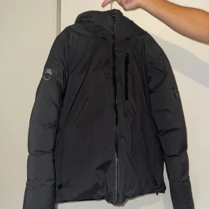 Svart dunjacka från Sail Racing L - Säljer en svart dunjacka från Sail Racing i storlek L. Jackan har huva, vattentäta dragkedjor och en bröstficka med Sail Racing-logga. Insidan har ett blått mönstrat foder som ger en cool kontrast. 