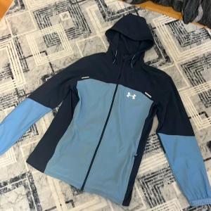 Blå vindjacka från Under Armour - Snygg tvåfärgad vindjacka från Under Armour i mörkblått och ljusblått. Jackan har huva, hel dragkedja framtill och logga på bröstet samt stor logotyp på ryggen. Perfekt för träning utomhus och har en sportig vibe.