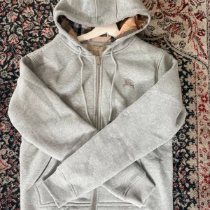 Burberry Zip Hoodie - Snygg grå hoodie från Burberry med dragkedja framtill, snörning i huvan och klassiskt rutigt foder. Broderad logga på bröstet och ribbade muddar. Perfekt för en chill och stilren look. Knappt använt, alla tags finns och kan skickas med!