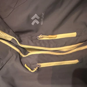 Grön skidjacka från 157 Funk, strl 160 - Snygg grön skidjacka från 157 Funk med gula dragkedjor och reflexdetaljer på ärmarna. Jackan har huva, flera fickor med dragkedja och är perfekt för blåsiga dagar. Materialet är slitstarkt och jackan har en sportig look.