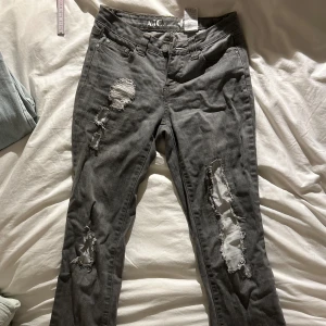 Grå slitna jeans från AJC, stl 34 - Grå jeans från AJC med coola slitningar och trasiga detaljer på benen. Klassisk femficksmodell med dragkedja och knapp. Jeansen har en rak passform och är tillverkade i bomull med lite stretch för extra komfort. Midjemått rakt över: 36cm och innerbenslängden är: 79cm