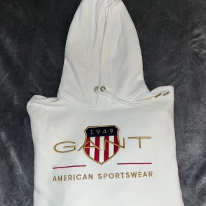 Vit hoodie från GANT med brodyr - Vit hoodie från GANT med broderad logga och sköld på bröstet. Klassisk huva med snörning och snygga gulddetaljer. Mjuk och skön bomullskvalitet, perfekt för en sportig och stilren look. Köpt från Johnells. Originalpris: 1200kr