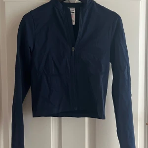 Marinblå zip-tröja från Gymshark XS - Säljer en marinblå långärmad zip-tröja från Gymshark i storlek XS. Tröjan är croppad, har dragkedja framtill och är tillverkad i ett stretchigt material som passar perfekt till träning. Snygg och stilren design med Gymshark-logga på ryggen.