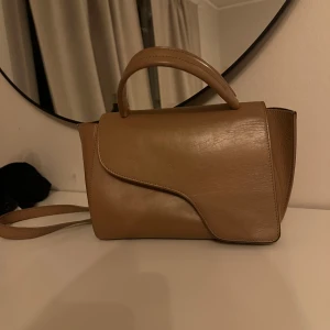 Beige/brun atp väska i skinn med handtag - Snygg beige axelväska i skinn med stilren design och avtagbar axelrem. Väskan har ett topphandtag och unikt format lock. Perfekt för dig som vill ha en trendig och praktisk väska med plats för det viktigaste. Knappen på ena sidan har ramlat av så man behöver sätta dit en ny knapp, så för tillfället går den inte att stänga. Nypris 5200kr