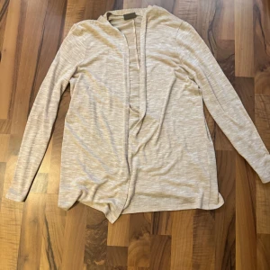 Beige kofta från Vero Moda, stl M - Ljusbeige, tunn och öppen kofta från Vero Moda i storlek M. Koftan har lång ärm och en enkel, stilren design utan knappar eller dragkedja. Perfekt att slänga över en t-shirt eller topp för en chill look.