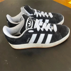 Adidas Campus svarta sneakers mocka - Svarta Adidas Campus sneakers i mocka med klassiska vita ränder och vit sula. Skorna har breda vita snören och en gummisula med fiskbensmönster. Ikonisk Adidas-logga på plösen och insidan. Perfekta för en avslappnad och stilren look.