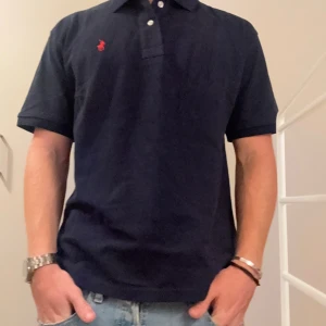Mörkblå piké från Polo Ralph Lauren - Klassisk mörkblå pikétröja från Polo Ralph Lauren i bomull. Tröjan har korta ärmar, krage med knappar och den ikoniska röda broderade loggan på bröstet. Classic fit-modell som passar till jeans eller shorts.