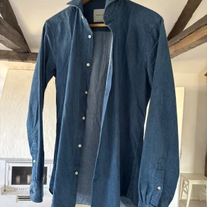 Blå skjorta från A Day's March - Klassisk blå jeansliknande skjorta från A Day's March i mjuk bomull. Skjortan har lång ärm, vit knappstängning och enkel krage. Perfekt för en clean och stilren look. Passar dig som gillar minimalistisk stil och vill ha något som funkar till många outfits. 