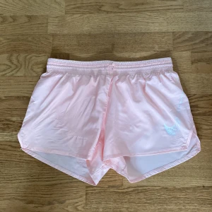 Ljusrosa träningsshorts från Nike - Snygga ljusrosa träningsshorts från Nike med vitt swoosh-tryck på ena benet. De har elastisk midja med snörning och är tillverkade i ett glansigt, lätt syntetmaterial. Perfekta för sport eller gym och har en loose passform för extra rörelsefrihet.
