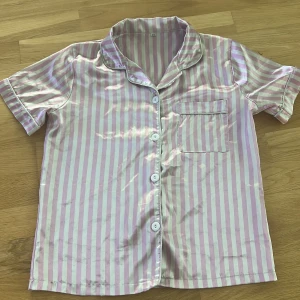 Rosa och vit randig pyjamasskjorta - Snygg pyjamasskjorta med rosa och vita ränder, kort ärm och klassisk krage. Skjortan har knappar framtill och en bröstficka. Materialet är glansigt och känns lyxigt mot huden, perfekt för chillkvällar hemma.