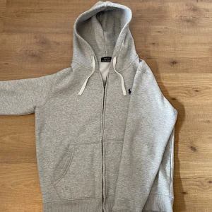 Grå hoodie från Polo Ralph Lauren - Snygg ljusgrå hoodie från Polo Ralph Lauren med dragkedja, huva med vita snören och klassisk broderad logga på bröstet. Mjuk bomullsblandning och känguruficka framtill. Perfekt för chill dagar eller när du vill ha en clean look.