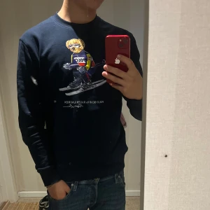 Mörkblå sweatshirt Polo Ralph Lauren - Säljer en mörkblå sweatshirt från Polo Ralph Lauren med ikoniska Polo Bear-trycket på bröstet. Tröjan har rund halsringning och långa ärmar, perfekt för en chill och stilren look. Passar dig som gillar streetwear och klassiska märken.