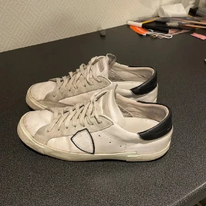 Philippe Model skor | 41  - Snygga vita sneakers från Philippe Model Paris med svarta detaljer på hälen och grå mockadetaljer. Skorna har snörning, rund tå och är tillverkade i skinn med en klassisk låg profil. Perfekta för dig som gillar stilrena och trendiga sneakers. Svagaste delen på skon är undersidan som är lite skadad men som ja inte tycker är något jätte stort problem då ja tidigare använt skorna. Pris går alltid att diskutera och så (Tveka inte att höra av dig😉)