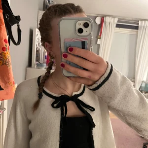 Vit kofta med svarta detaljer från Nelly - Supersöt vit kofta från Nelly i storlek XXS med svarta kanter och rosett vid halsen. Långärmad och knäppning framtill, perfekt att slänga över ett linne. Mjuk och stickad känsla, passar till många outfits.