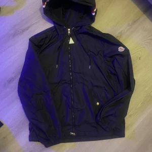 Svart vindjacka från Moncler med huva - Snygg svart vindjacka från Moncler med huva och dragkedja framtill. Jackan har Moncler-logga på ärmen, snörning vid huvan och en cool detalj med rödvit rand på huvkanten. Perfekt för dig som vill ha en stilren och sportig look.