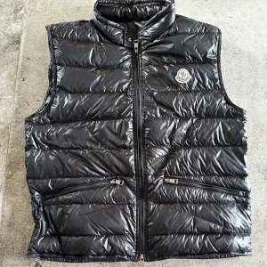 Svart dunväst från Moncler stl 2 - Snygg svart dunväst från Moncler med glansig finish och klassisk logga på bröstet. Västen har hög krage, två dragkedjefickor framtill och stängs med dragkedja. Perfekt för lager-på-lager och ger en clean look. Fint använt skick. 