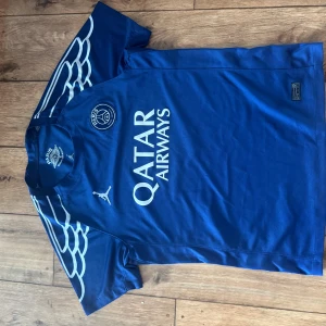 Blå PSG x Jordan fotbollströja 2025 - Snygg blå PSG fotbollströja från samarbetet med Jordan. Varför inte bära en vinnares gestalt med Champions league vinnarna 2025. Perfekt street wear passar och med den vita detaljen kan man ha på sig den med vad som helst.