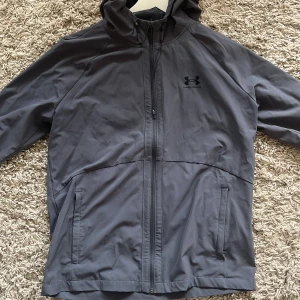 Grå vindjacka från Under Armour - Säljer en grå vindjacka från Under Armour med huva och dragkedja hela vägen. Jackan har meshfoder, två sidofickor och en stilren look. Perfekt för blåsiga dagar och enkel att slänga på sig när du är på språng.