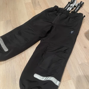 Svarta täckbyxor med reflexdetaljer - Svarta täckbyxor från Polarn O. Pyret med elastisk midja, justerbara hängslen och reflexdetaljer på benen. Perfekta för kalla dagar och utomhusaktiviteter. Byxorna har förstärkta knän och resår vid benslut för extra komfort. Jätte fint skick på dom! Storlek 104!