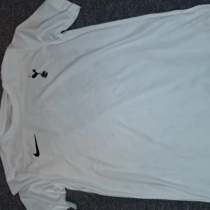 Vit Tottenham Hotspur Nike fotbollströja - Säljer en vit Tottenham Hotspur fotbollströja från Nike med Dri-FIT-material som andas. Tröjan har korta ärmar, svart Nike-logga och Spurs-emblem på bröstet. Perfekt för träning eller match, lätt och bekväm passform.