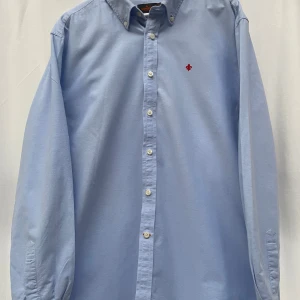 Ljusblå skjorta Morris 2XL - Snygg ljusblå skjorta från Morris i storlek 2XL med klassisk button-down krage och vita knappar. Broderad röd logga på bröstet ger en clean och stilren vibe. Tillverkad i mjuk bomull som känns skön mot huden. Perfekt för dig som gillar enkel och tidlös stil. Nypris 1399kr
