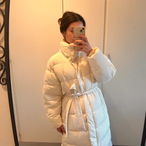 Beige vinter pufferjacka från BikBok XS - Säljer en beige pufferjacka från BikBok i storlek XS. Jackan är quiltad, har bälte i midjan och dragkedja med tryckknappar. Storlek 34 men är oversized så skulle även passa dig som har storlek 36-38. Jackan har använts 1 gång men måste tyvärr säljas pga stor storlek.  —-NOTERA: lång vinterjacka som täcker enda till under knäna. 