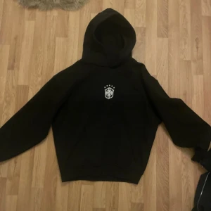 Svart hoodie Brasilien hoodie från Wunna - Svart hoodie med vit logga på bröstet och stor huva. Klassisk modell med långa ärmar och ribbade muddar. Perfekt för chill dagar eller när du vill ha en enkel och stilren look.