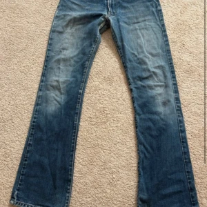Blå bootcut jeans med slitningar - Snygga blå jeans med bootcut passform och slitna detaljer på benen. Jeansen har klassisk femficksdesign och är tillverkade i ett robust denimtyg. Perfekta för dig som gillar en avslappnad och trendig stil.