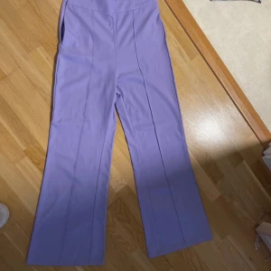 Lila vida byxor med pressveck - Snygga lila byxor med vida ben och markerade pressveck framtill. Byxorna har hög midja och är tillverkade i ett mjukt, vävt material som ger en clean look. Perfekta för dig som vill sticka ut med färg och stil.