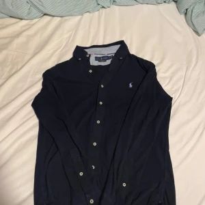 Mörkblå skjorta från Ralph Lauren - Snygg mörkblå skjorta från Ralph Lauren i featherweight mesh. Klassisk button-down krage, vita knappar och ikoniska broderade loggan på bröstet. Perfekt för dig som gillar stilrena och tidlösa plagg med en sportig touch.