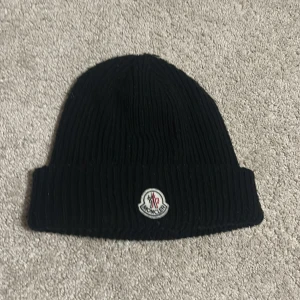 Svart ribbstickad mössa från Moncler - Svart ribbstickad mössa från Moncler med klassisk uppvikt kant och broderad logotyp framtill. Tillverkad i mjukt material som håller dig varm under kyliga dagar. Enkel och stilren design som passar till det mesta. Nfc funkar ej