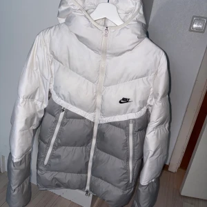 Nike pufferjacka i vitt och grått - Snygg pufferjacka från Nike i vitt och ljusgrått med huva och dragkedja framtill. Jackan har två snedställda fickor med dragkedja och klassisk Nike-logga på bröstet. Perfekt för kalla dagar och riktigt skön att bära.