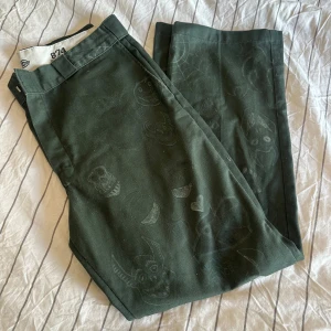 Mörkgröna Dickies 874 byxor med tryck - Säljer ett par mörkgröna Dickies 874 Original Fit byxor med unika tryck av olika tecknade ansikten och motiv. Byxorna har raka ben, klassisk passform och är tillverkade i slitstarkt material. Perfekta för dig som vill sticka ut med en cool streetstyle.