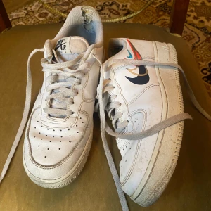 Nike Air Force 1 vita sneakers - Nike Air Force 1 sneakers i vitt skinn med klassisk rund tå och platt sula. Skorna har snörning och detaljer med färgglada swoosh-loggor i blått, rött och guld på sidan. Perforeringar på tån för extra ventilation. Perfekt för dig som gillar streetstyle.