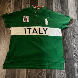 Grön pikétröja Polo Ralph Lauren Italy - Grön pikétröja från Polo Ralph Lauren med vit broderad logga och stor text 'ITALY' över bröstet. Snygga detaljer som broderat märke på vänster sida och krage med rödvit rand. Klassisk passform och tillverkad i bomull.