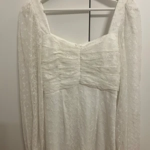 Vit knälång klänning från Bubbleroom - Superfin vit knälång klänning från Bubbleroom med transparenta långärmar och broderade blommor över hela klänningen. Den har rynkade detaljer vid bysten och dragkedja i ryggen. Perfekt för dig som vill ha en romantisk och elegant look.