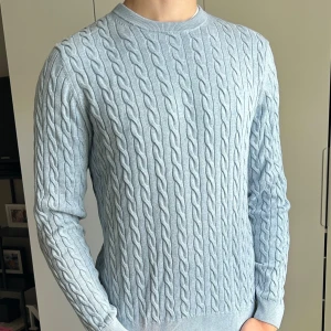 Ljusblå kabelstickad tröja - Säljer en ljusblå kabelstickad tröja med rund hals och lång ärm. Tröjan har ett klassiskt flätmönster över hela plagget och är tillverkad i ett mjukt stickat material. Perfekt för dig som gillar en stilren och fräsch look. Broderat ”Bondelid” på vänster bröst. Modellen är 190cm, 78kg. Skriv om ni har några funderingar:)