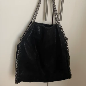Svart Stella McCartney liknande väska - Snygg svart axelväska med glansig finish och grova silverfärgade kedjor längs kanterna och som axelrem. Väskan är i ett jätte fint skick men kommer tyvärr inte till användning längre! Vid frågor eller pris så kontakta mig💕