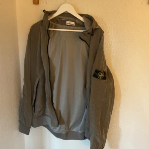 Grå vindjacka Stone Island XXL - Snygg grå vindjacka från Stone Island i storlek XXL. Jackan har huva, dragkedja framtill och den klassiska Stone Island-loggan på ärmen. Tillverkad i lätt polyester med meshfoder för extra komfort. Perfekt för dig som gillar streetwear och stilrena detaljer.