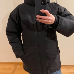 Svart vinterjacka från Marmot - Svart vinterjacka från Marmot med huva och dragkedja. Jackan är vadderad och har en klassisk, rak passform med stora fickor framtill. Perfekt för kalla dagar och har diskret Marmot-logga på bröstet. Snygg och funktionell design. Det står i jackan Storlek XXS vilket motsvarar 36. Bruksanvänd. Inga defekter. 