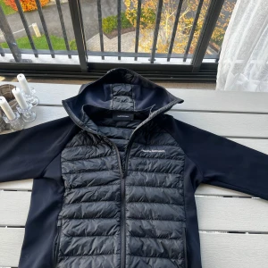 Svart pufferjacka Peak Performance M - Svart pufferjacka från Peak Performance i storlek M. Jackan har huva, quiltad framsida och dragkedja hela vägen. Ärmar och sidor är i slät polyester, medan framsidan är vadderad. Snygg logga på bröstet och ärmen. Perfekt för kyliga dagar.