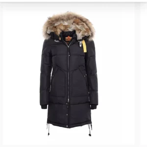 Svart Parajumpers dunjacka med päls - Säljer en svart lång dunjacka från Parajumpers med fluffig päls runt huvan. Jackan har dragkedja framtill, snörning vid nederkant och långa ärmar. Den har en gul detalj vid halsen och är perfekt för kalla vinterdagar. Snygg och varm design med coola detaljer.